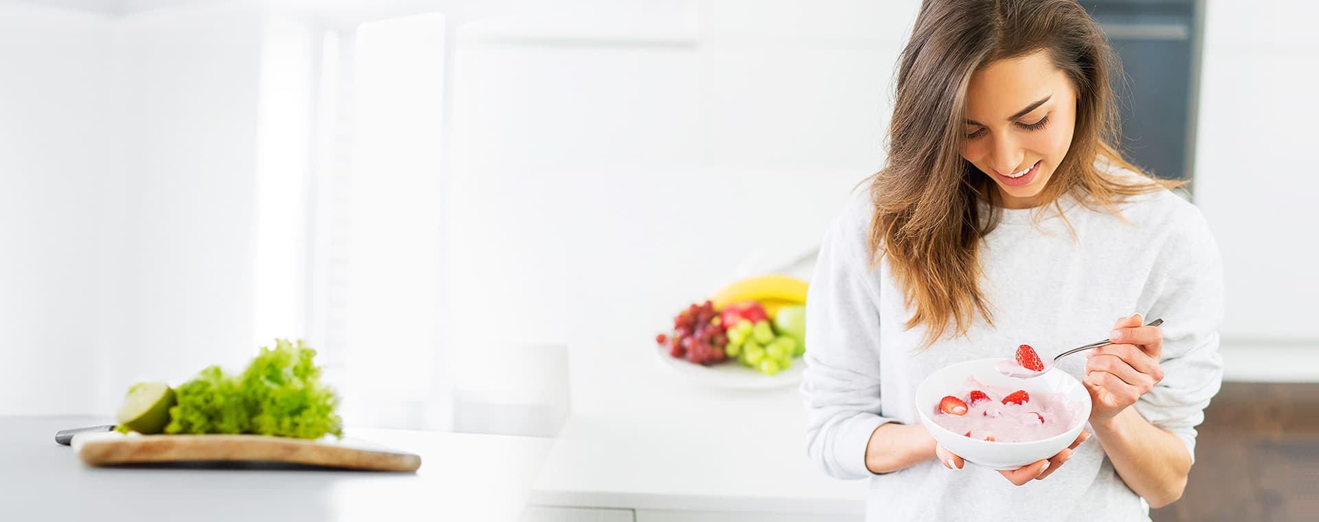 Woman in a bright kitchen eating a bowl of yoghurt with strawberries, fresh fruit in the background